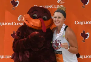 A smiling runner poses with a turkey mascot, both wearing race medals.