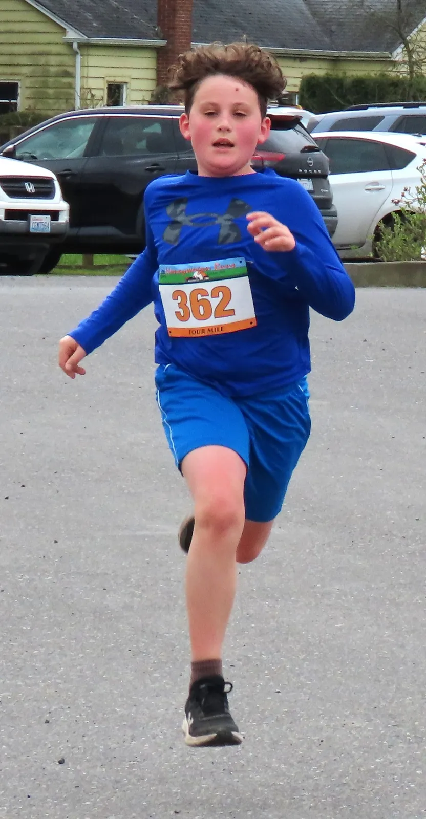 A young runner with a bib number 362 runs on a paved surface, wearing a blue long-sleeved shirt and blue shorts.