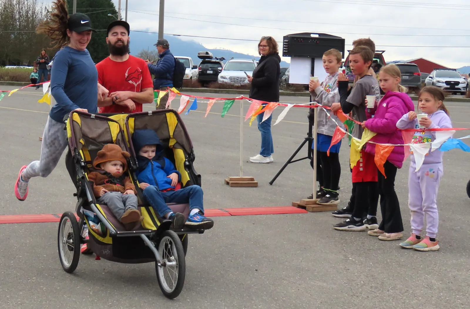 A woman pushes a double stroller with two children across a finish line while a crowd of children and adults watch.