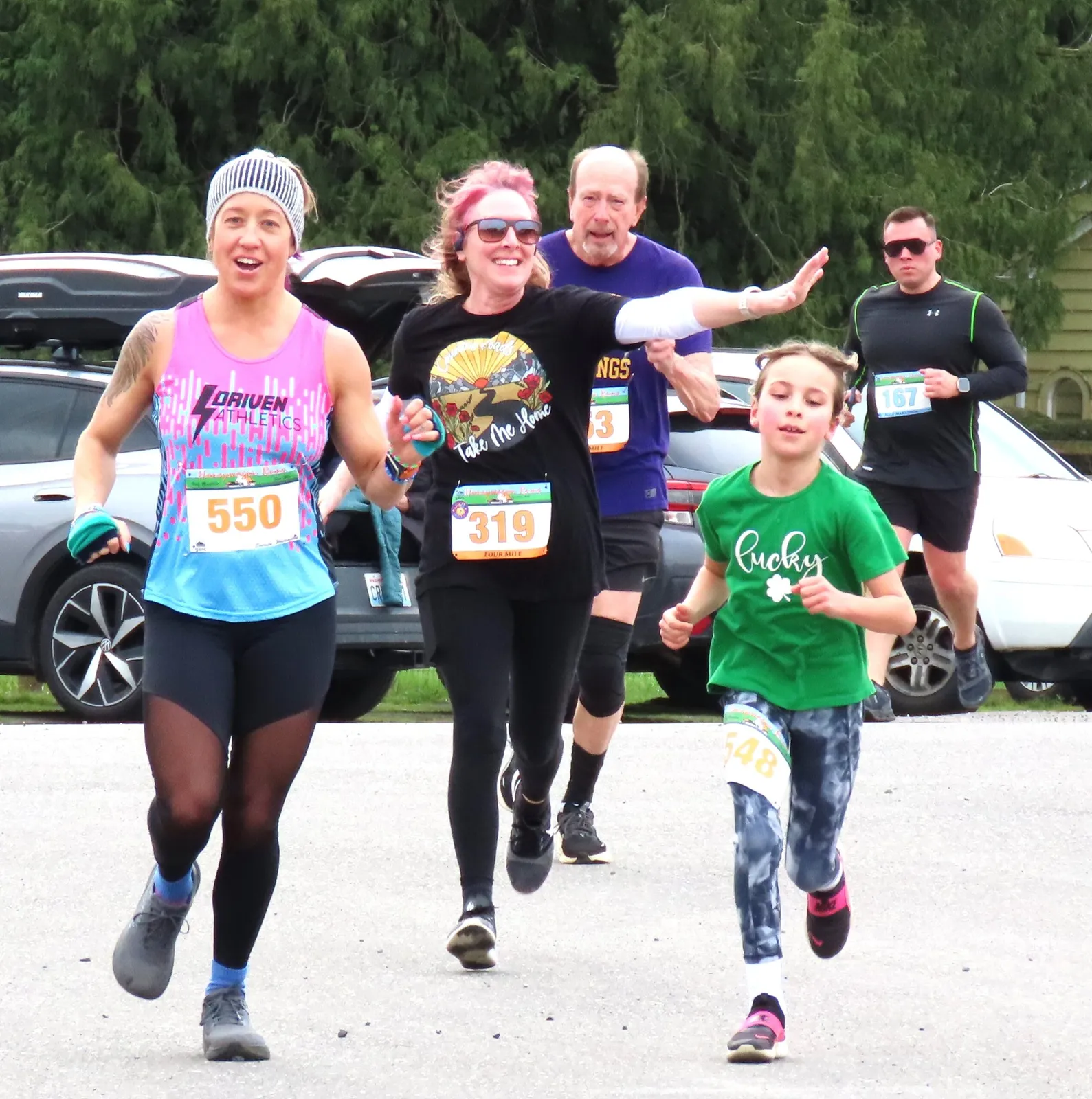 A group of runners, including a child, crossing the finish line with smiles and waves.