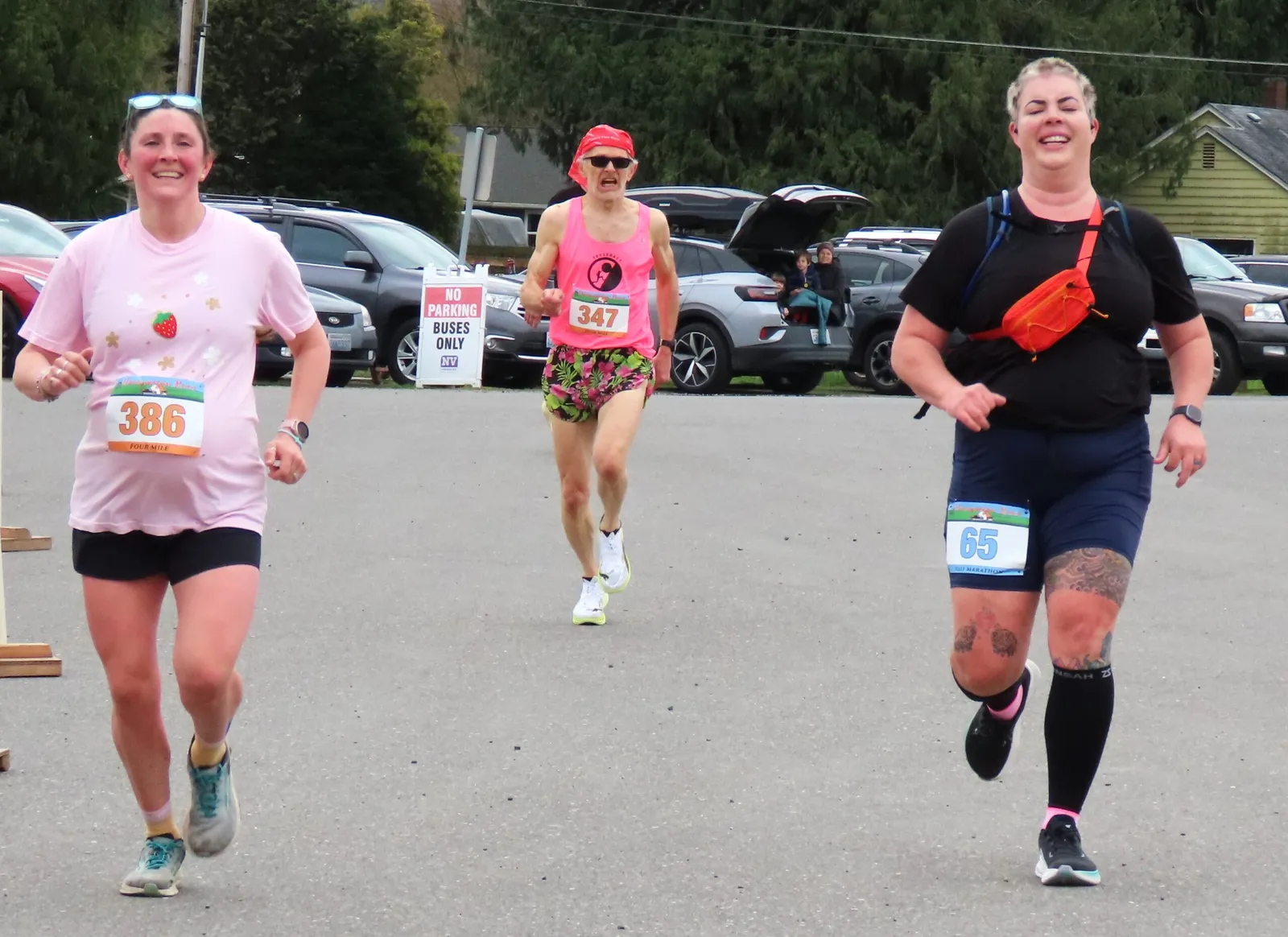 Three runners are shown from the waist down, running towards the camera. The runner on the left wears a pink shirt with a strawberry design and bib number 386. The runner in the middle wears a pink tank top with bib number 347 and floral shorts. The runner on the right wears a black shirt and dark shorts with bib number 65.