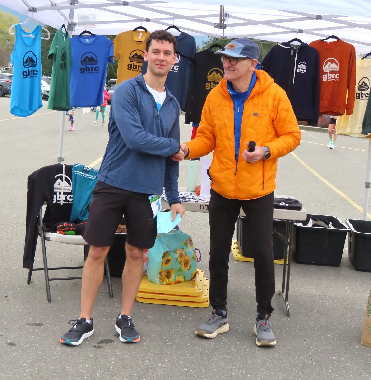 Two men shaking hands in front of a tent with race merchandise.