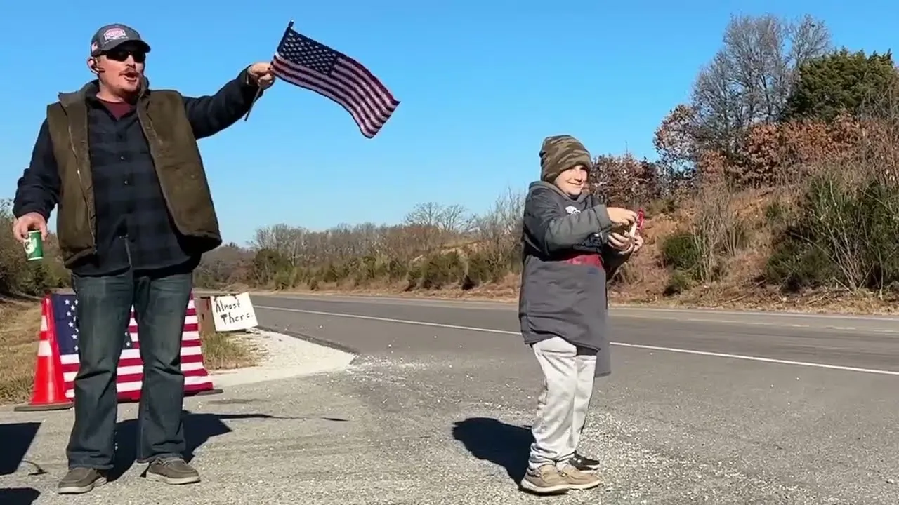 A man waves an American flag and a child holds a small item, standing by the side of a road with a sign that says 'Almost There'.