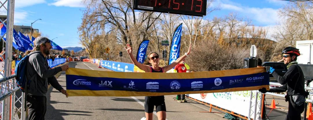 A female runner crosses the finish line with her arms raised in celebration, holding a banner with race sponsors.