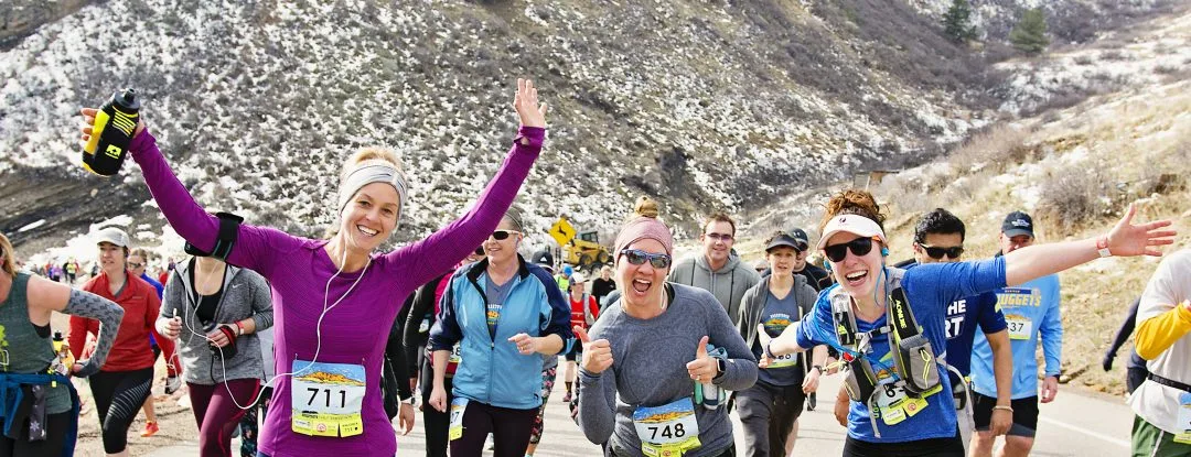 A group of happy runners participating in a half marathon, with some waving and smiling at the camera, set against a scenic, mountainous backdrop with patches of snow.