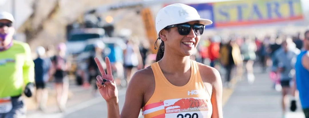 A smiling female runner wearing a white hat and sunglasses gives a peace sign at the start line of the Horsetooth Half Marathon.