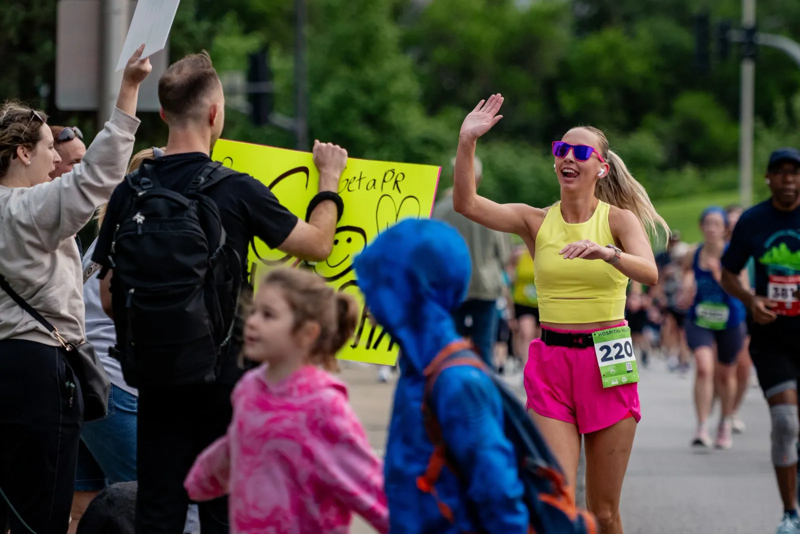 A female runner in a yellow top and pink shorts waves to spectators while running in a race, with a sign that says 'Get a PR' visible.