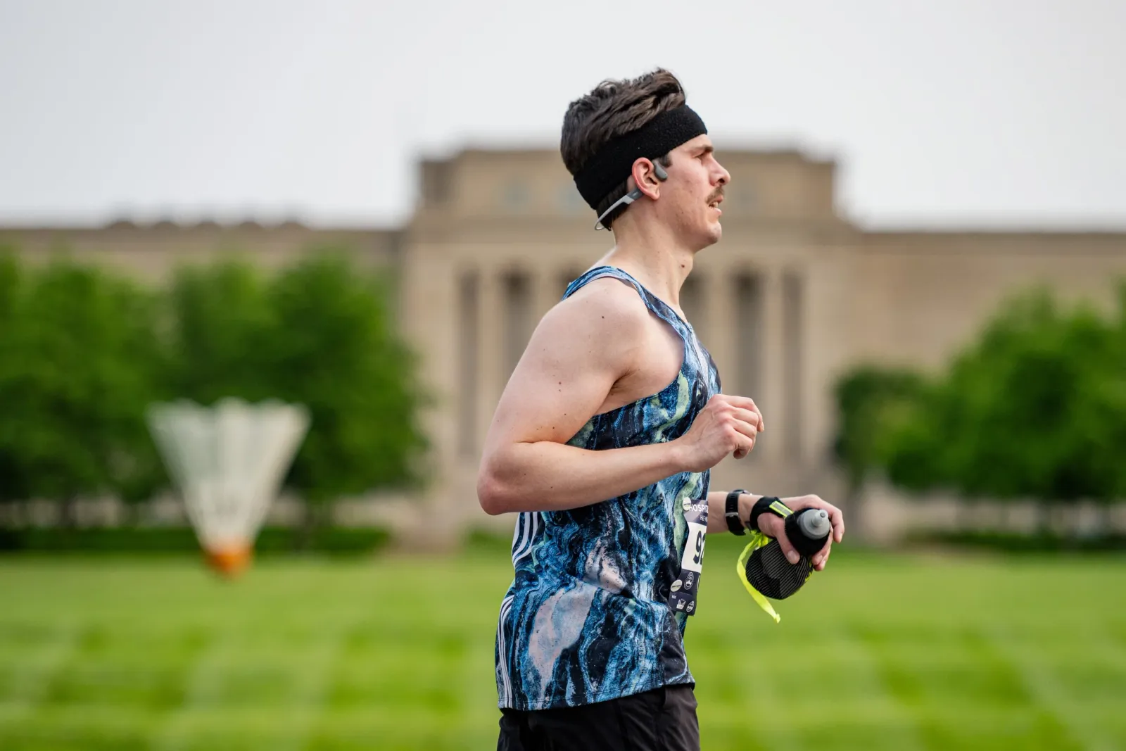 A male runner in a blue and black patterned tank top runs with a water bottle, wearing a headband and earbuds, with a building and trees in the background.