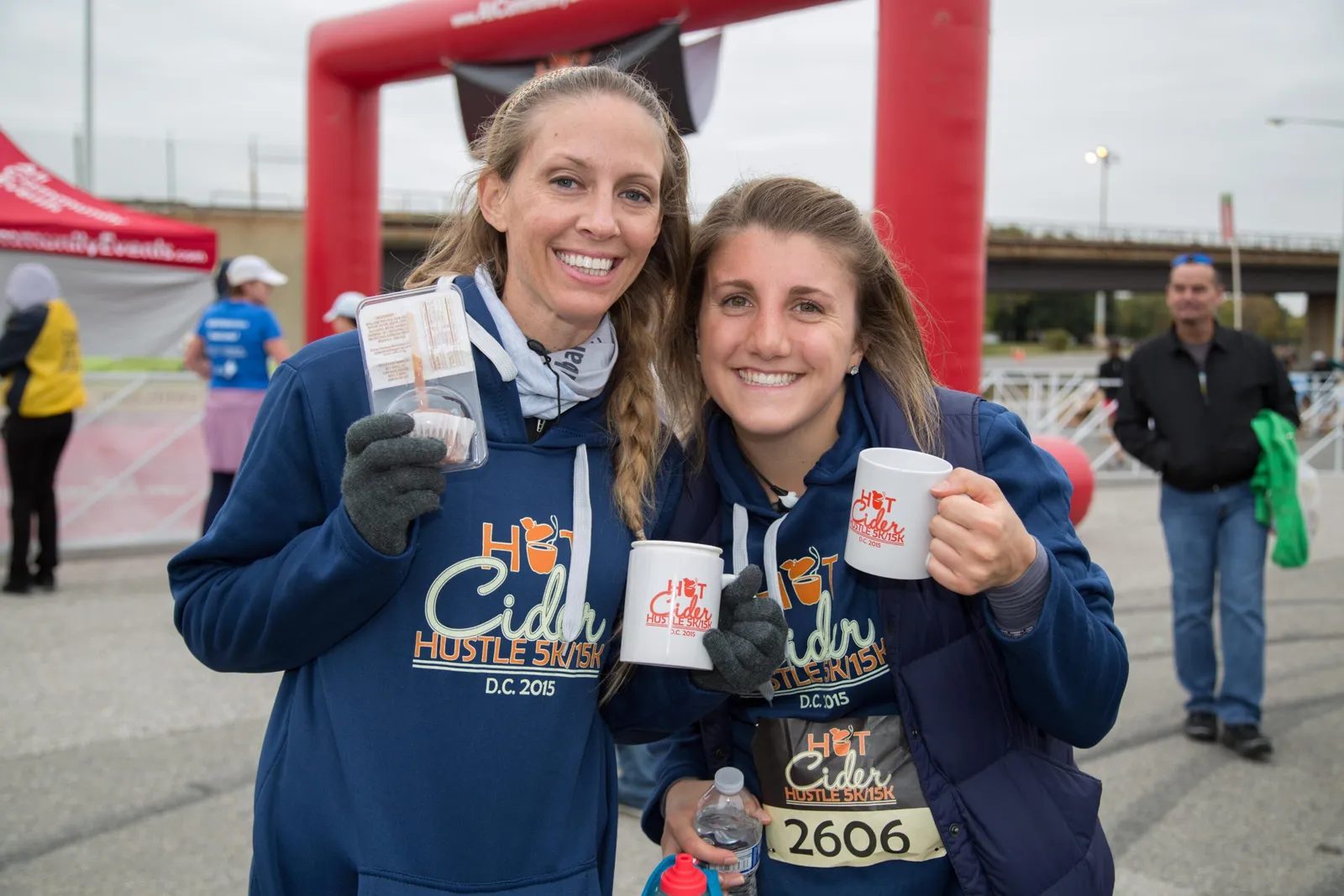 Two smiling women at the Hot Cider Hustle race, one holding a cupcake and the other a mug, with a red inflatable arch in the background.