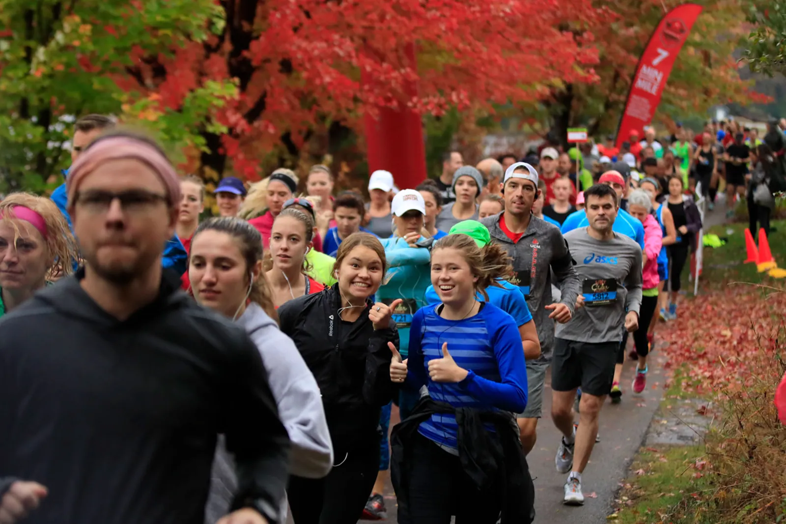 A group of runners, some smiling and giving thumbs up, run along a tree-lined path with red autumn leaves.