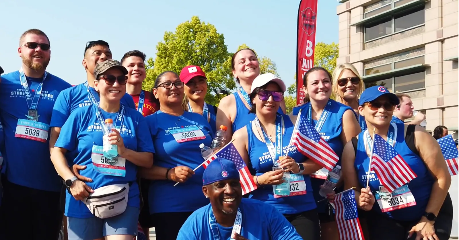 A group of smiling runners wearing blue race t-shirts and medals, some holding American flags, pose for a photo after completing the Stars and Stripes Half Marathon.