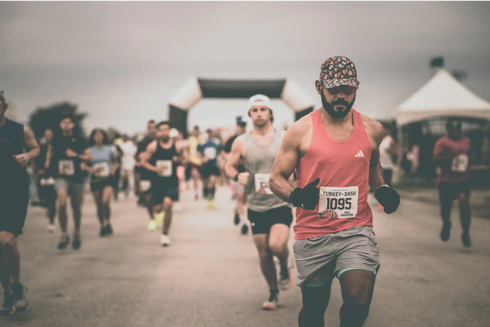 A runner wearing a pink tank top and a patterned cap runs towards the camera, with other runners and a start/finish arch in the background.