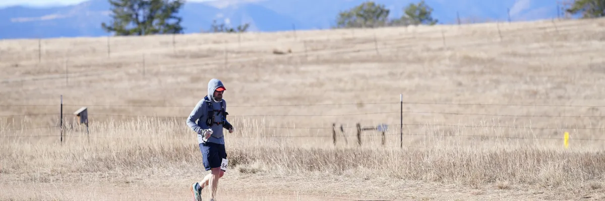 A male runner with a beard and a red hat runs on a dirt trail through a dry, grassy field with distant mountains under a blue sky.