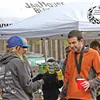 Two runners, one wearing an orange jacket and a medal, chat near a tent.