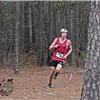 A runner wearing a red tank top and black shorts runs on a dirt trail through a pine forest.