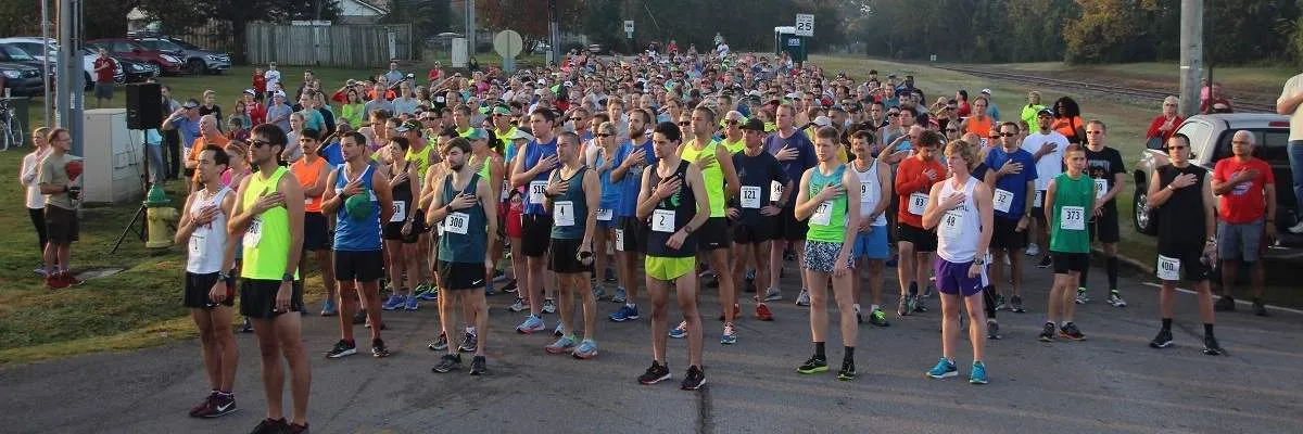 A large group of runners stand in rows at the start line of a race, with their hands over their hearts.