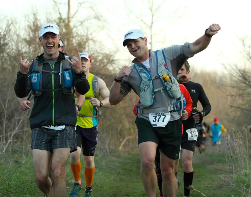 Two runners with excited expressions running on a trail, one giving a rock-on hand gesture and the other with a fist pump.