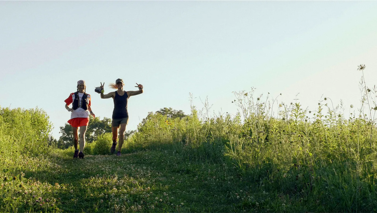 Two female runners smiling and giving peace signs while running up a grassy hill on a trail.