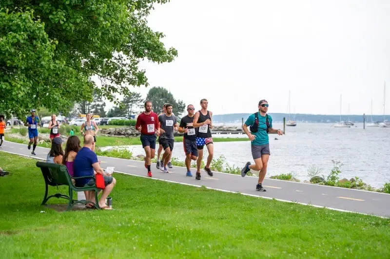 Runners on a paved path next to a body of water with sailboats in the background.