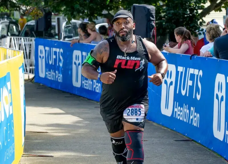 A male runner wearing a black tank top with 'Black Men RUN' logo, bib number 2885, and knee braces, running on a road with spectators and sponsor banners in the background.