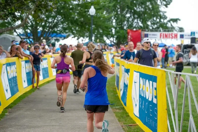 Runners approaching the finish line at the Independence Rhode Race, with spectators cheering from behind a yellow banner.