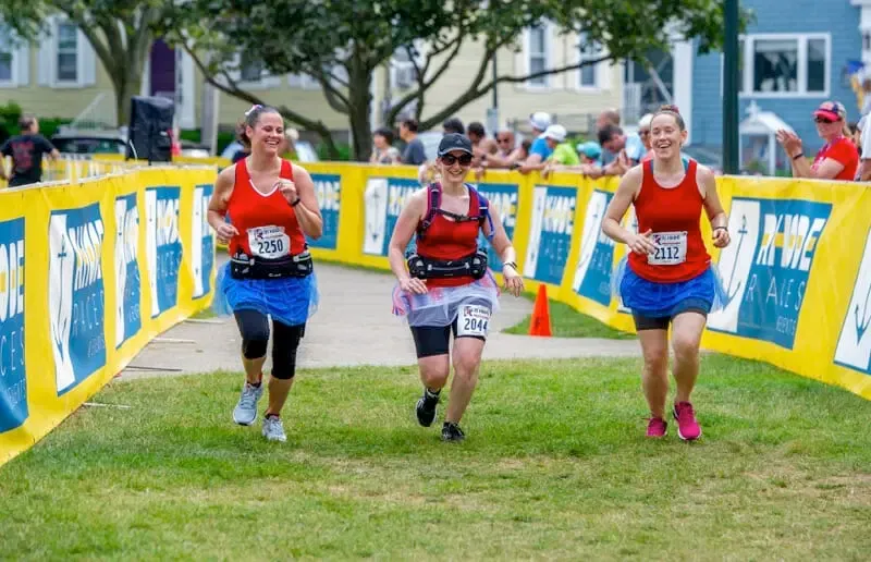 Three women in red tank tops and blue tutus running towards the finish line, smiling.