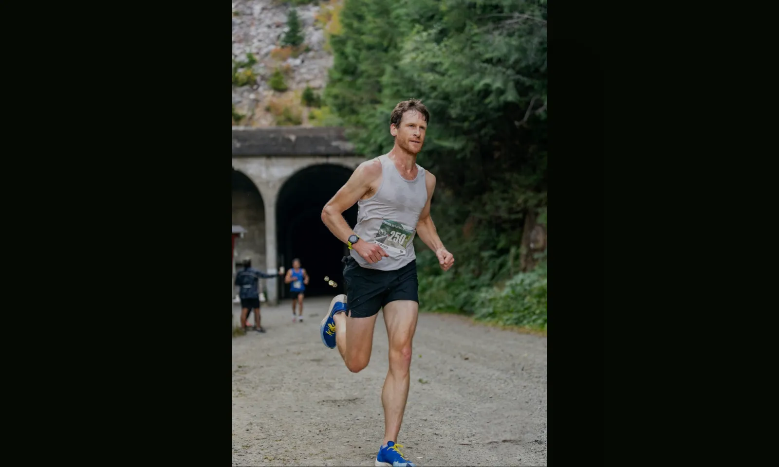 A male runner in a grey tank top and black shorts runs on a dirt path with a tunnel in the background.