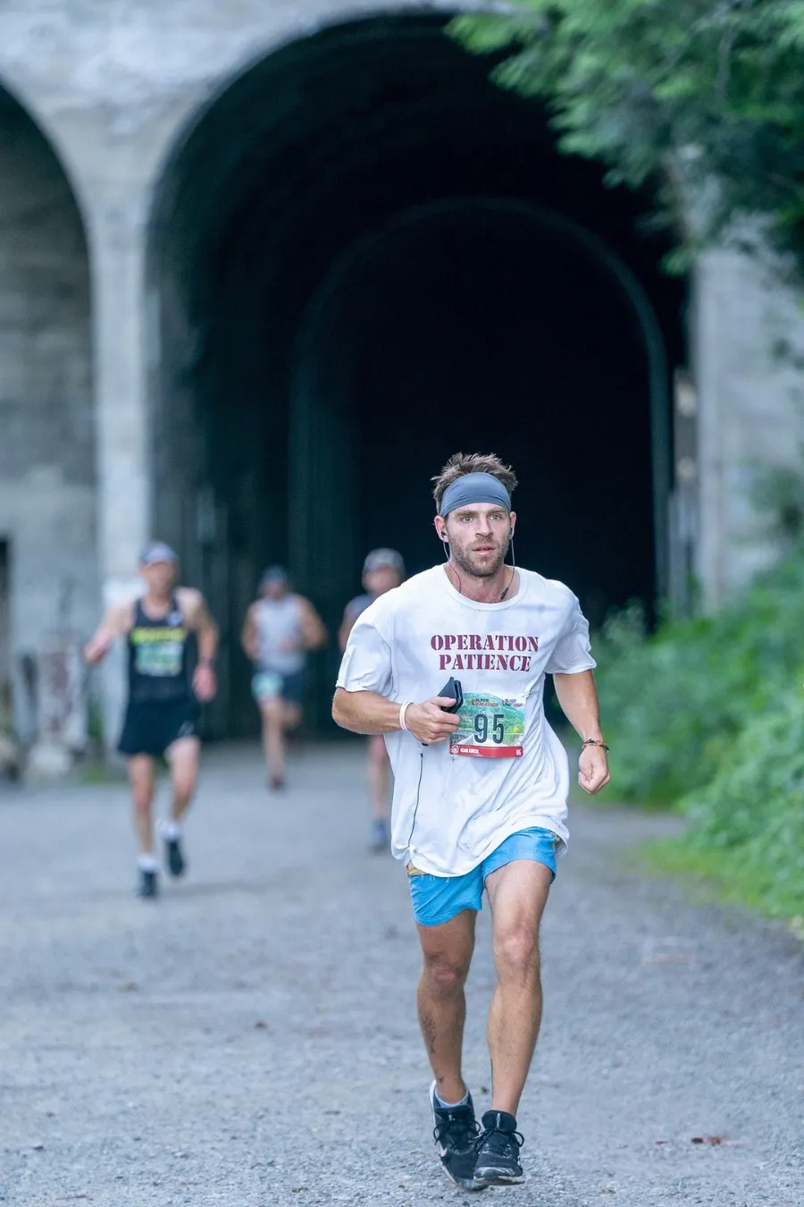 A male runner wearing a white t-shirt with 'OPERATION PATIENCE' and bib number 95, runs towards the camera exiting a dark tunnel, with other runners in the background.