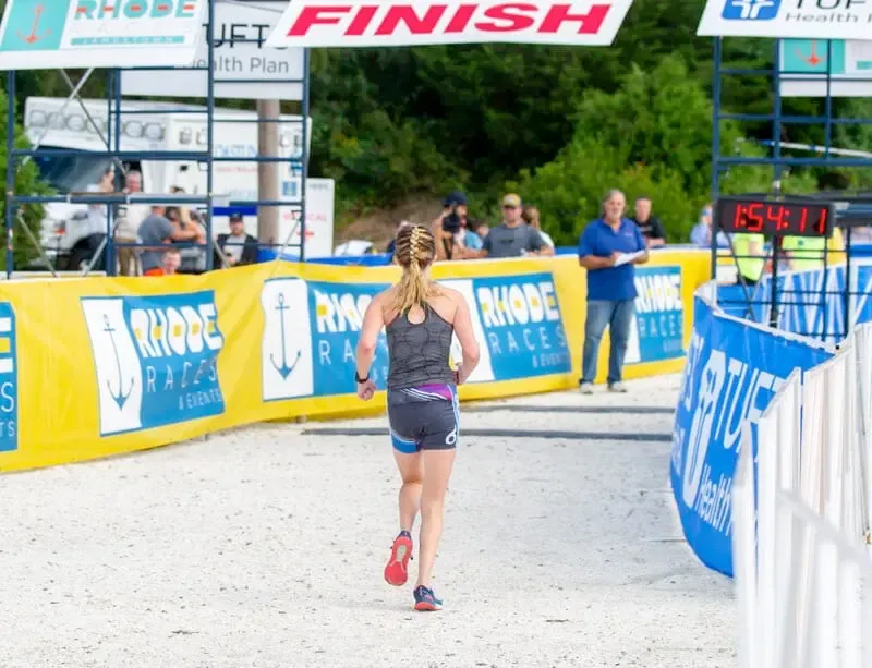 A female runner approaches the finish line, with race banners and a timing clock visible.