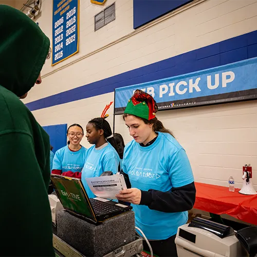 Volunteers in blue t-shirts assist runners at a bib pickup station, with a banner reading 'BIB PICK UP' in the background.