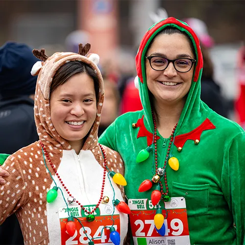 Two women smiling at the camera, one dressed as a reindeer and the other as an elf, wearing festive necklaces with Christmas lights.
