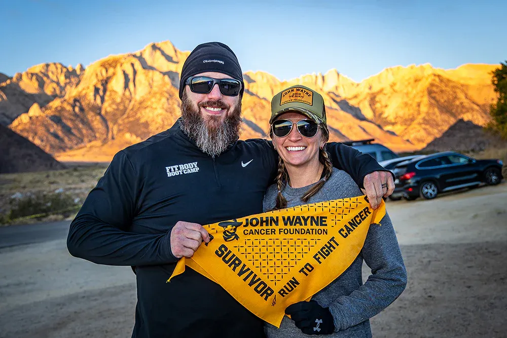 A man and a woman smiling at the camera, holding a yellow banner that says 'John Wayne Cancer Foundation Survivor Run to Fight Cancer'.