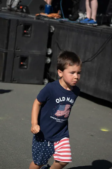 A young boy in a navy blue t-shirt and patriotic shorts runs forward with a determined expression.