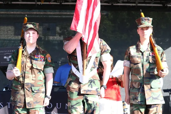 Three individuals in camouflage uniforms stand at attention, holding rifles and an American flag, on a stage.