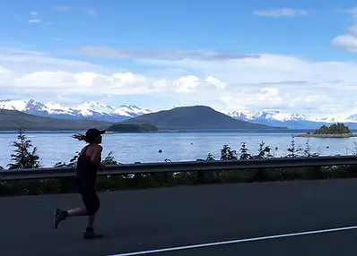 A runner runs along a road with a scenic view of a bay and snow-capped mountains in the background.