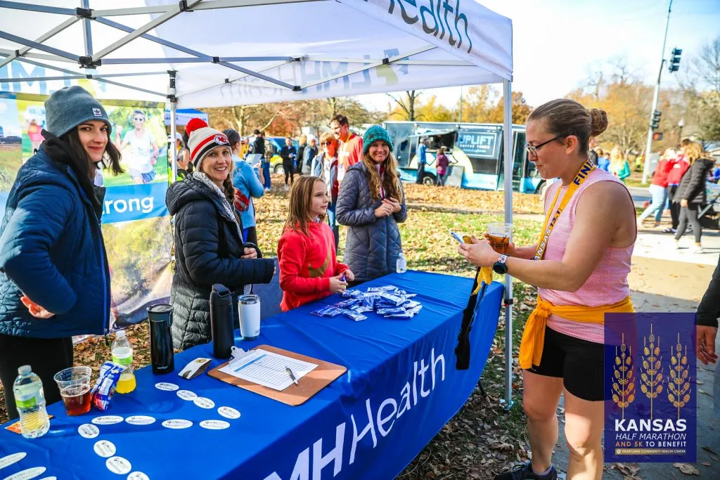 People at a race event booth, with a runner wearing a medal and holding a drink.