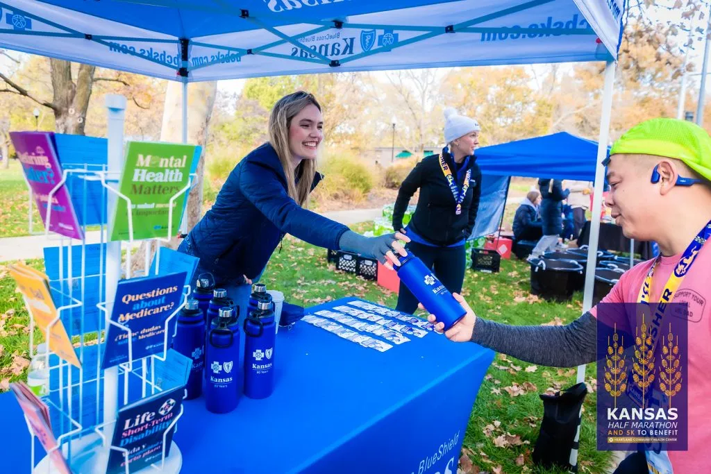 A volunteer hands a blue water bottle to a runner at a Kansas Half Marathon booth.