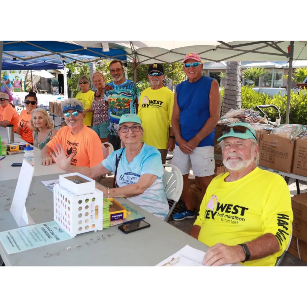 A group of people, some wearing race-themed shirts, are gathered around a table, possibly at a race registration or information booth.