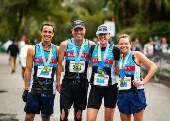 Four runners, two men and two women, wearing blue and black race singlets and medals, smiling at the camera after completing the Kiawah Marathon.