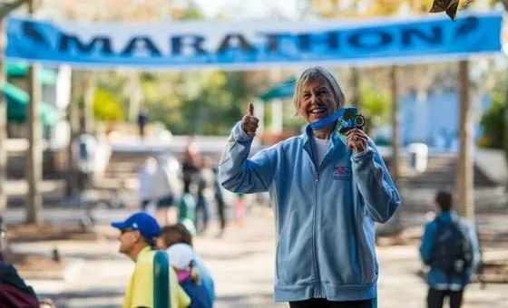A smiling woman gives a thumbs up and shows off her marathon medal under a blue 'MARATHON' banner.