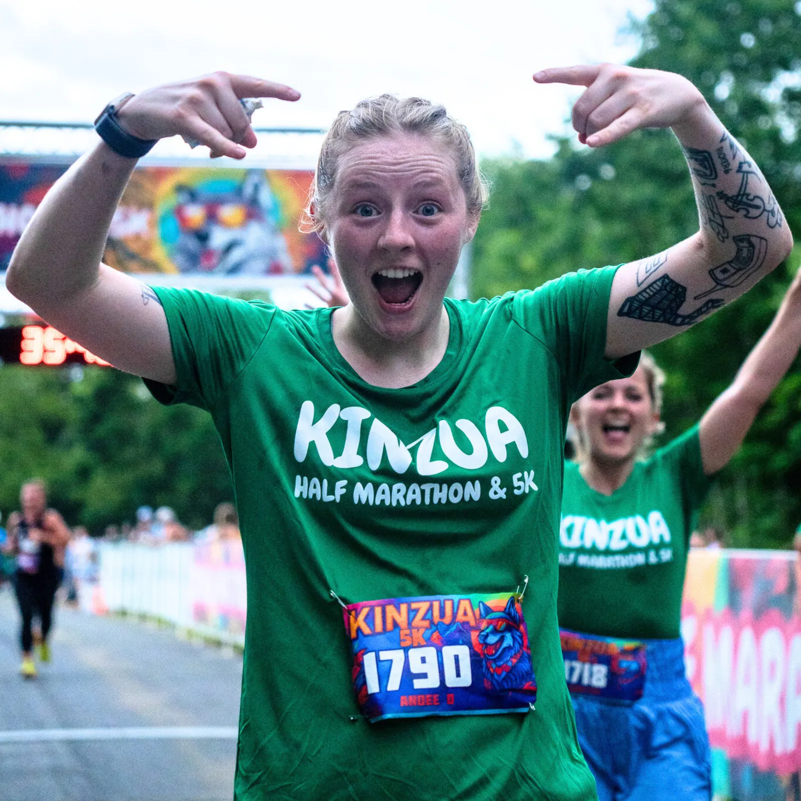 A jubilant female runner wearing a green Kinzua Half Marathon & 5K shirt crosses the finish line, pointing to herself with an excited expression. Another runner is visible behind her, also celebrating.