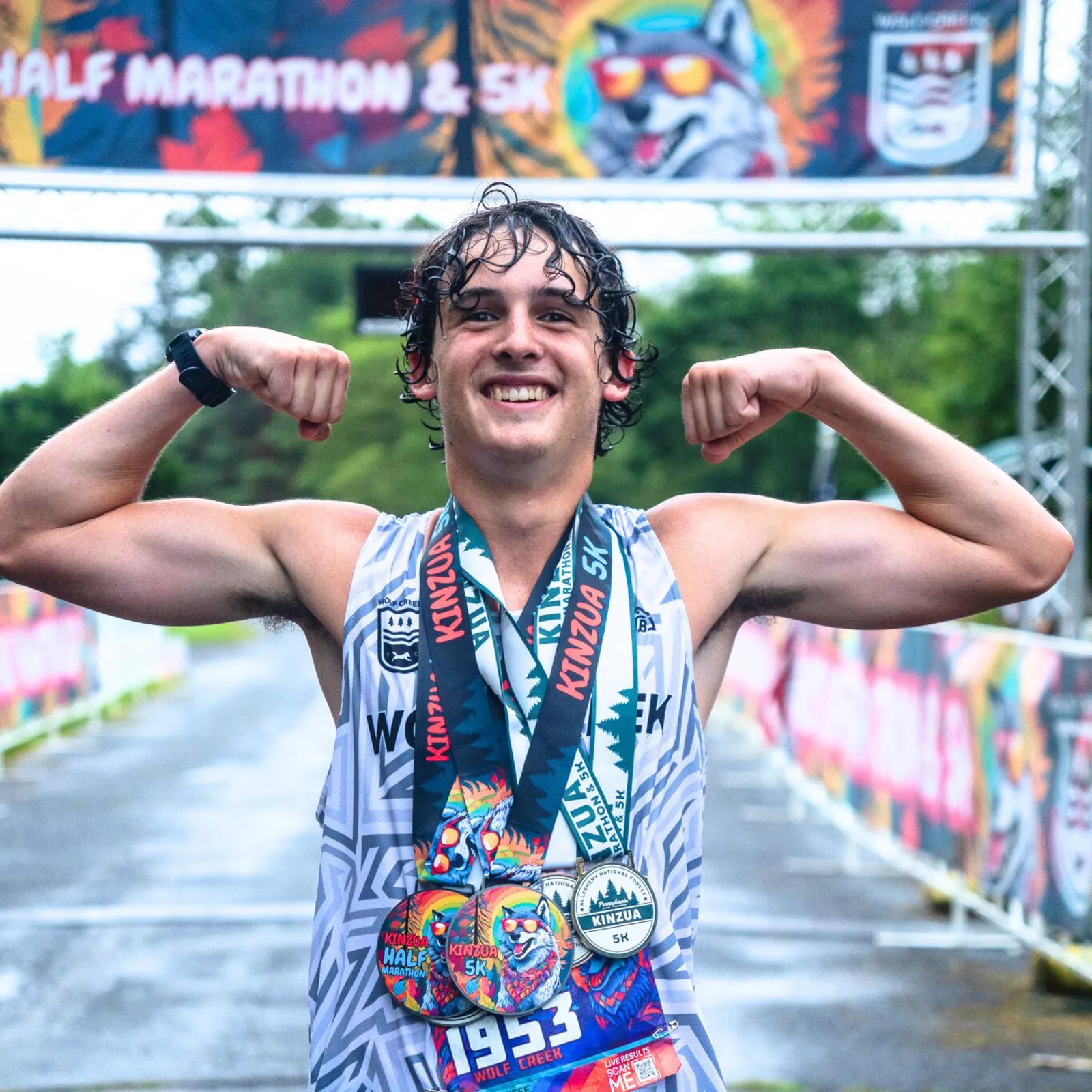 A young man with wet hair smiles and flexes his biceps, wearing multiple race medals around his neck, standing at a finish line with a race banner in the background.