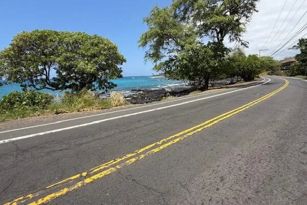 A scenic road curves alongside the ocean with lush trees on the left and a glimpse of buildings on the right.