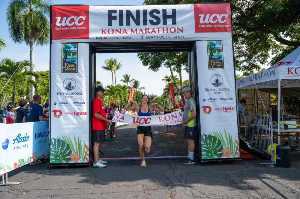 A female runner joyfully breaks the finish line tape at the Kona Marathon, flexing her muscles as two race officials hold the banner.