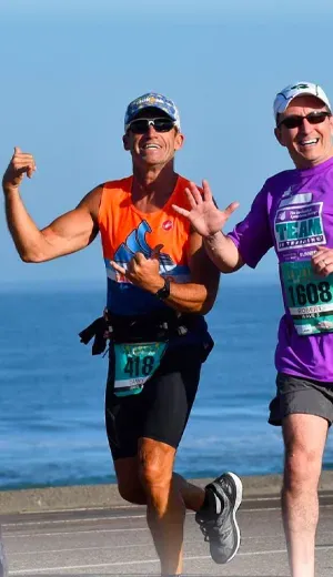 Two male runners smiling and running along a road with the ocean in the background.