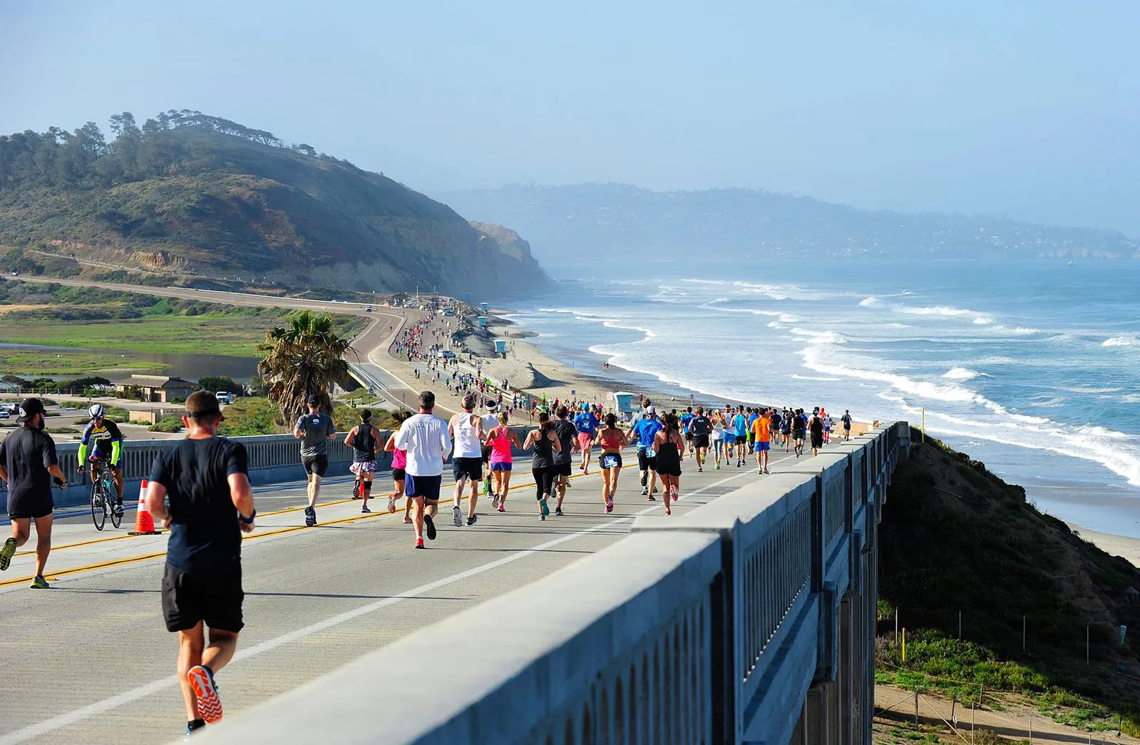 Runners race along a bridge with the ocean and coastline visible in the background.