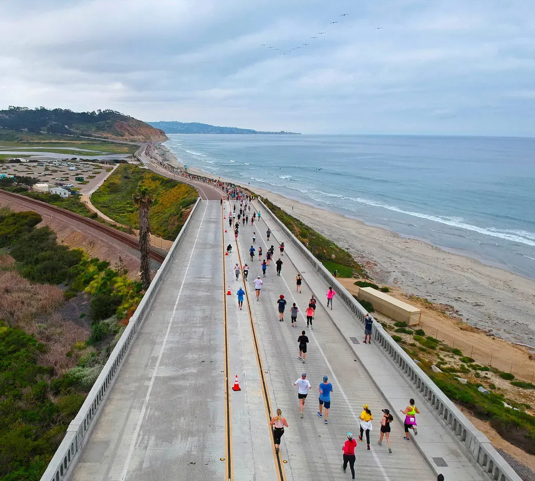 Runners race along a coastal highway with the ocean on one side and hills on the other.