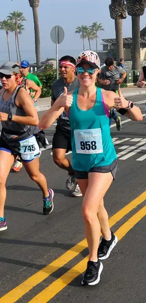 A female runner with bib number 958 gives a thumbs up while running in a race, with palm trees and the ocean in the background.