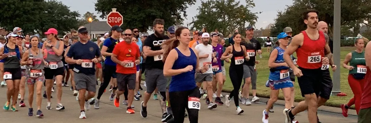 A diverse group of runners are starting a race, with bib numbers visible on their chests. The sky is overcast, and trees and houses line the street.