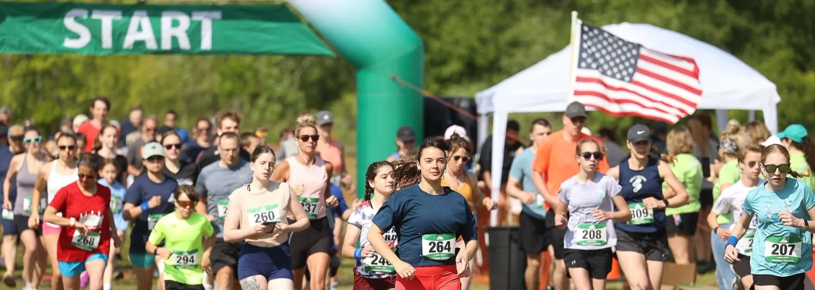A large group of runners are starting a race, with a green 'START' banner overhead and an American flag visible in the background.